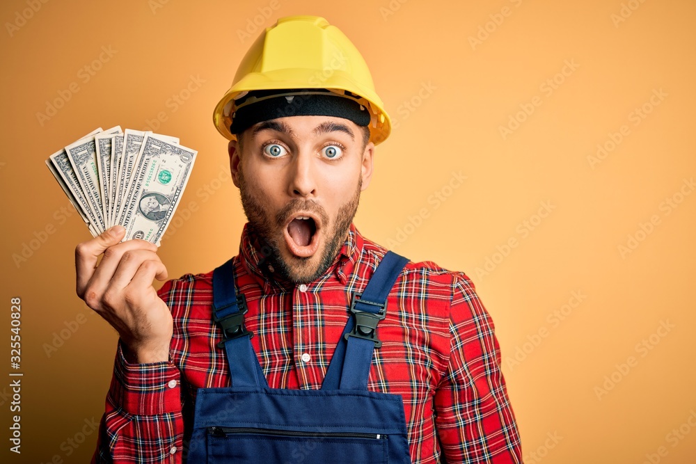 Young builder man wearing safety helmet holding dollars as payment over yellow background scared in shock with a surprise face, afraid and excited with fear expression Young builder man wearing safety helmet holding dollars as payment over yellow background scared in shock with a surprise face, afraid and excited with fear expression