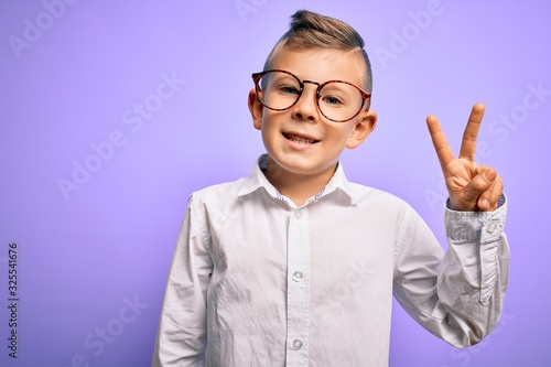 Young little caucasian kid with blue eyes wearing glasses and white shirt over purple background smiling with happy face winking at the camera doing victory sign. Number two.