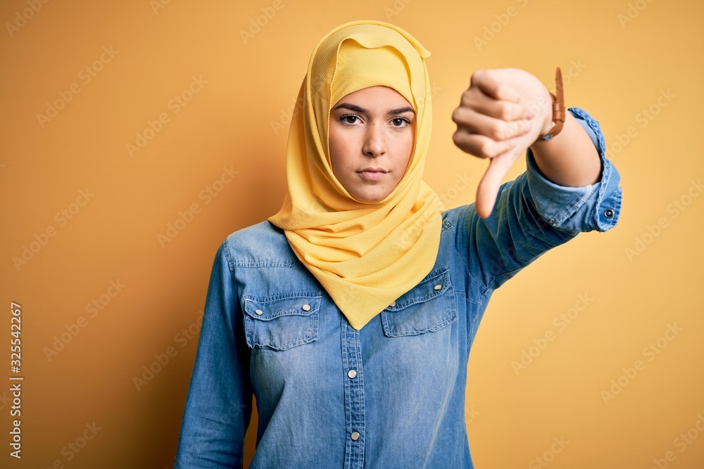 Young beautiful girl wearing muslim hijab standing over isolated yellow background looking unhappy and angry showing rejection and negative with thumbs down gesture. Bad expression. Young beautiful girl wearing muslim hijab standing over isolated yellow background looking unhappy and angry showing rejection and negative with thumbs down gesture. Bad expression.