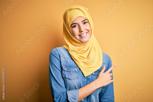 Young beautiful girl wearing muslim hijab standing over isolated yellow background cheerful with a smile of face pointing with hand and finger up to the side with happy and natural expression on face © Krakenimages.com