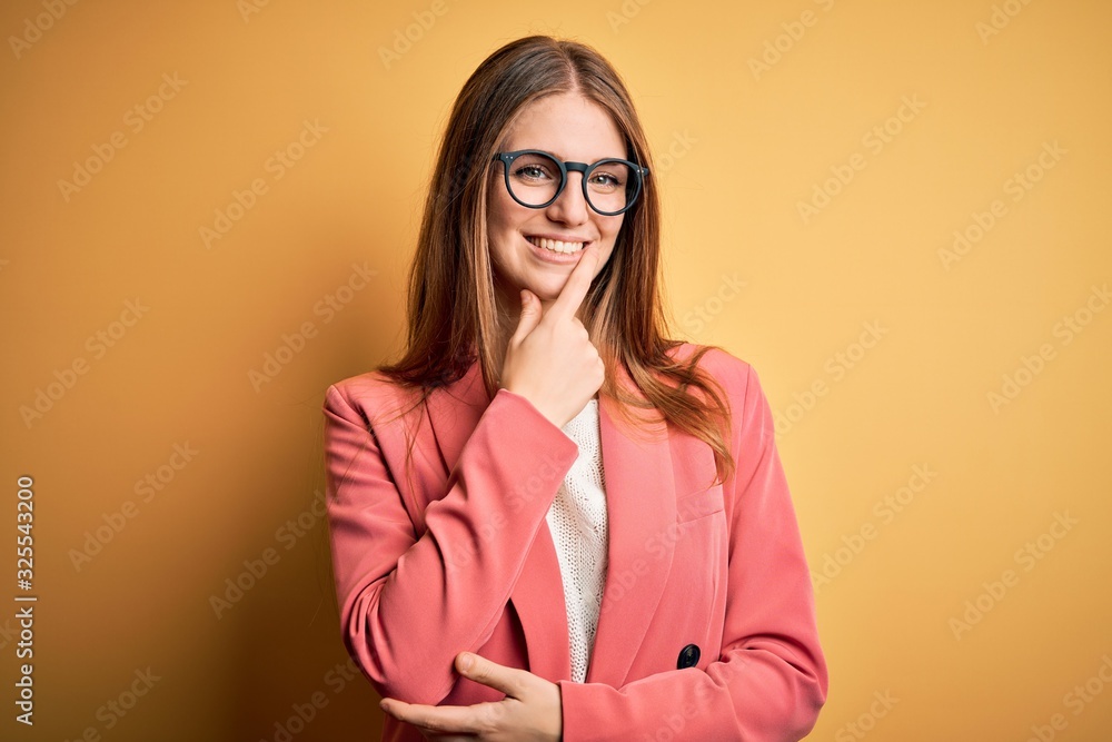 Young beautiful redhead woman wearing jacket and glasses over isolated yellow background looking confident at the camera with smile with crossed arms and hand raised on chin. Thinking positive.