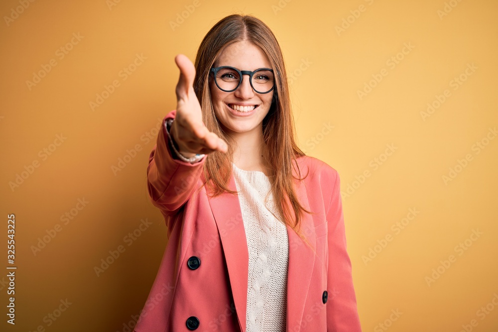 Young beautiful redhead woman wearing jacket and glasses over isolated yellow background smiling friendly offering handshake as greeting and welcoming. Successful business.