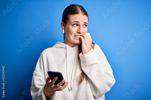 Young redhead sportswoman doing sport listening to music using earphones and smartphone looking stressed and nervous with hands on mouth biting nails. Anxiety problem.