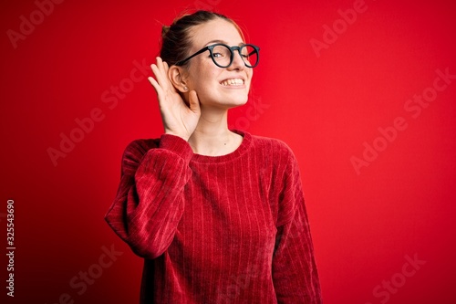 Young beautiful redhead woman wearing casual sweater over isolated red background smiling with hand over ear listening an hearing to rumor or gossip. Deafness concept.