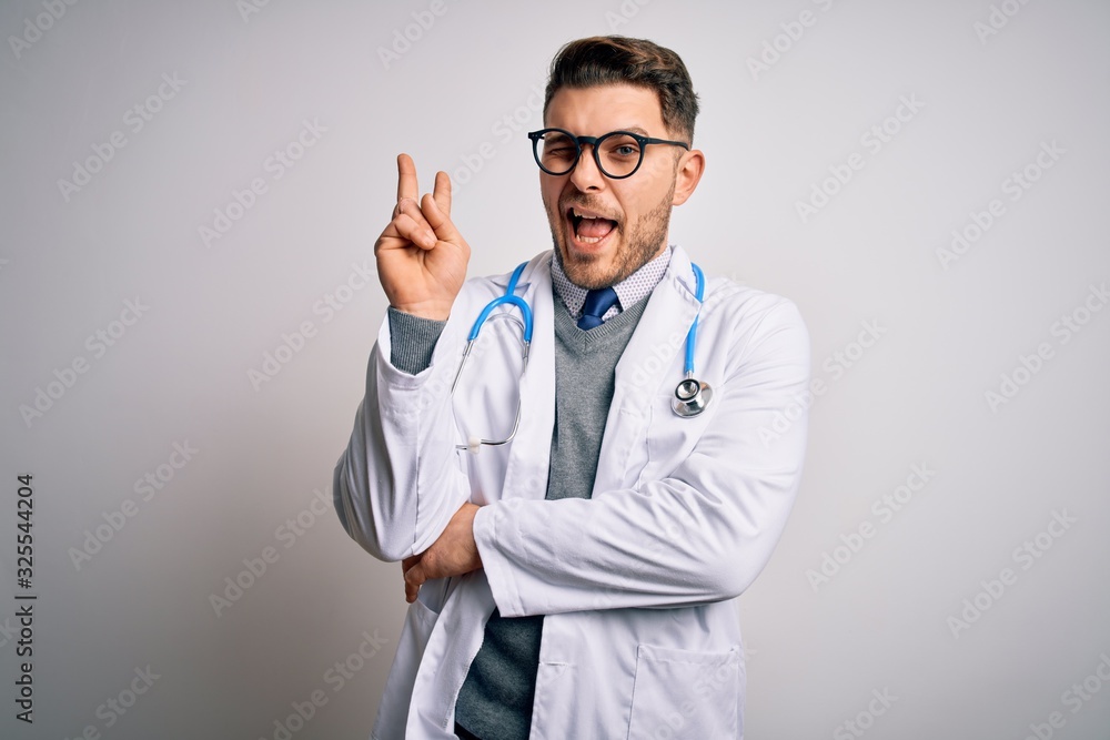 Young doctor man with blue eyes wearing medical coat and stethoscope over isolated background smiling with happy face winking at the camera doing victory sign with fingers. Number two. Young doctor man with blue eyes wearing medical coat and stethoscope over isolated background smiling with happy face winking at the camera doing victory sign with fingers. Number two.