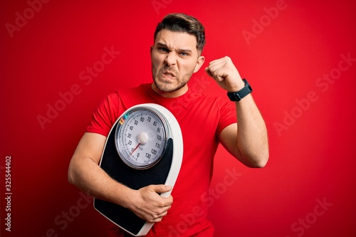 Young fitness man with blue eyes holding scale dieting for healthy weight over red background annoyed and frustrated shouting with anger, crazy and yelling with raised hand, anger concept