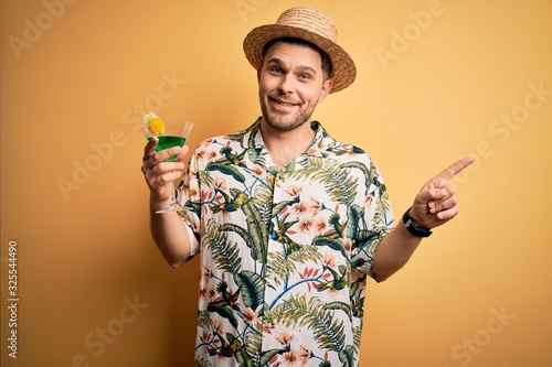 Billede på lærred Young man on vacation wearing summer hat drinking a party cocktail over yellow b