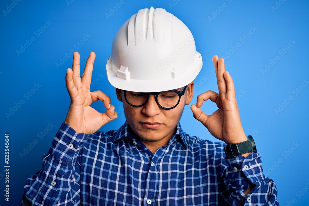 Young handsome engineer latin man wearing safety helmet over isolated blue background relax and smiling with eyes closed doing meditation gesture with fingers. Yoga concept.