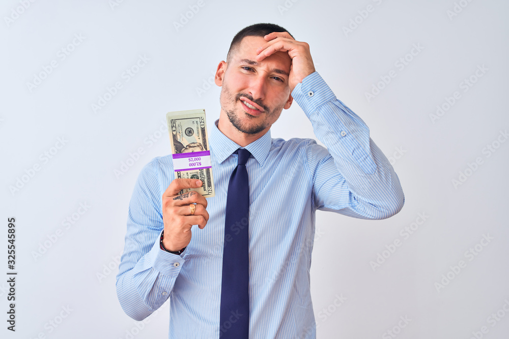 Young handsome business man holding a bunch of dollars bank notes over isolated background stressed with hand on head, shocked with shame and surprise face, angry and frustrated. Fear and upset Young handsome business man holding a bunch of dollars bank notes over isolated background stressed with hand on head, shocked with shame and surprise face, angry and frustrated. Fear and upset