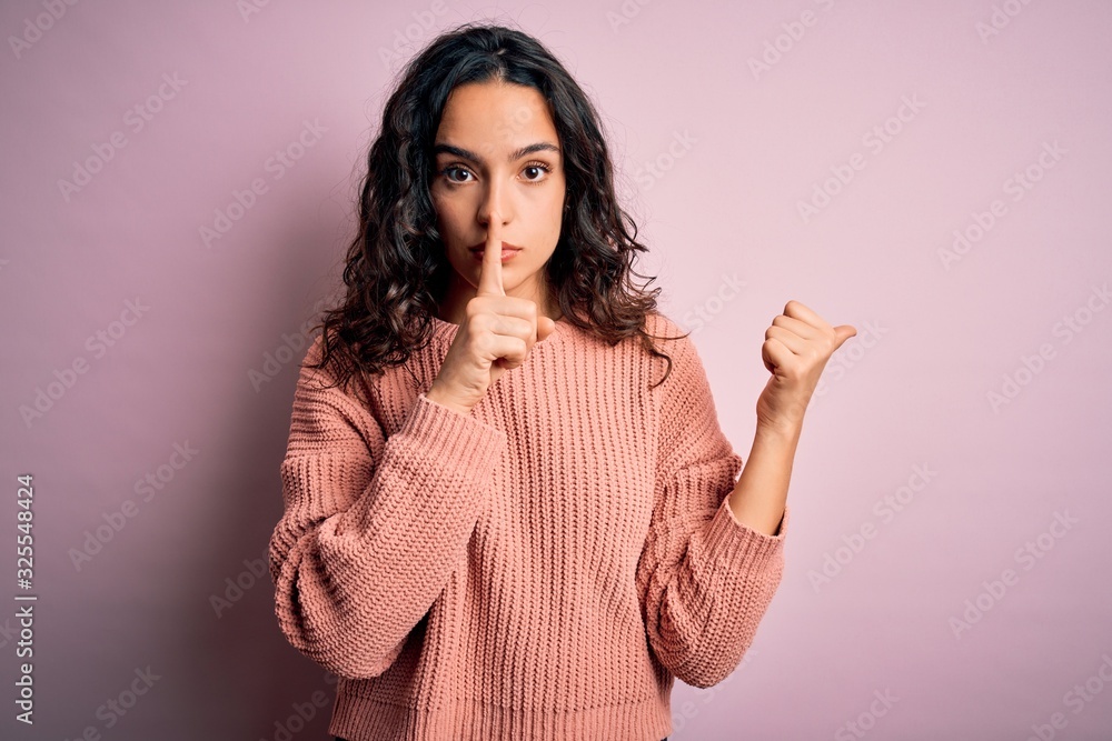 Young beautiful woman with curly hair wearing casual sweater over isolated pink background asking to be quiet with finger on lips pointing with hand to the side. Silence and secret concept. Young beautiful woman with curly hair wearing casual sweater over isolated pink background asking to be quiet with finger on lips pointing with hand to the side. Silence and secret concept.
