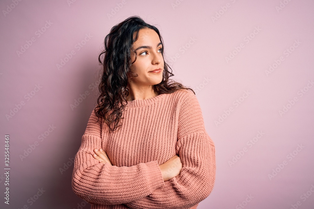 Young beautiful woman with curly hair wearing casual sweater over isolated pink background looking to the side with arms crossed convinced and confident Young beautiful woman with curly hair wearing casual sweater over isolated pink background looking to the side with arms crossed convinced and confident
