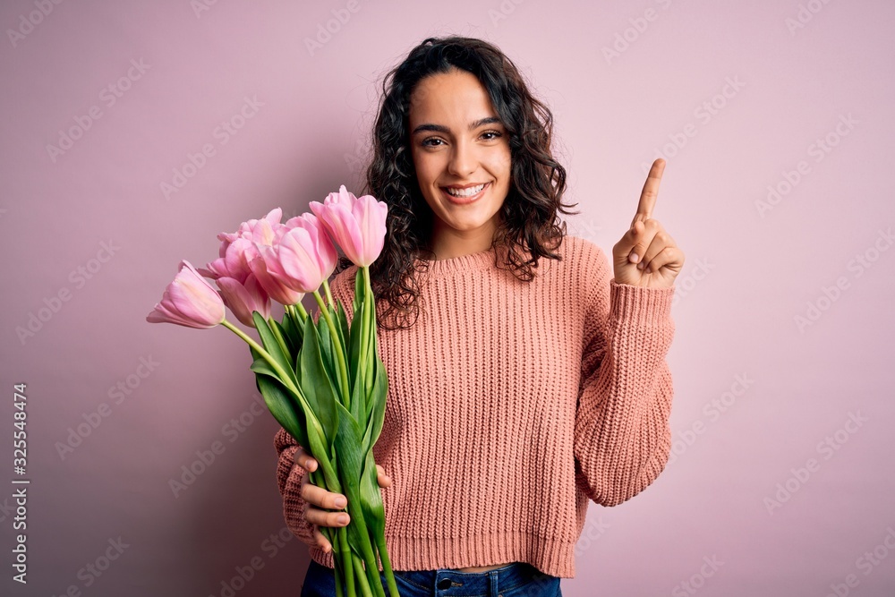 Young beautiful romantic woman with curly hair holding bouquet of pink tulips pointing finger up with successful idea. Exited and happy. Number one. Young beautiful romantic woman with curly hair holding bouquet of pink tulips pointing finger up with successful idea. Exited and happy. Number one.