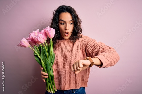 Young beautiful romantic woman with curly hair holding bouquet of pink tulips Looking at the watch time worried, afraid of getting late