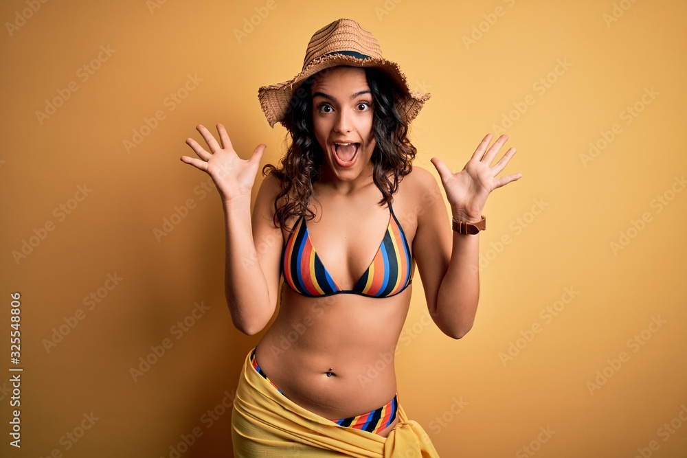 Young beautiful woman with curly hair on vacation wearing bikini and summer hat celebrating crazy and amazed for success with arms raised and open eyes screaming excited. Winner concept Young beautiful woman with curly hair on vacation wearing bikini and summer hat celebrating crazy and amazed for success with arms raised and open eyes screaming excited. Winner concept