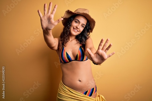 Young beautiful woman with curly hair on vacation wearing bikini and summer hat showing and pointing up with fingers number ten while smiling confident and happy.