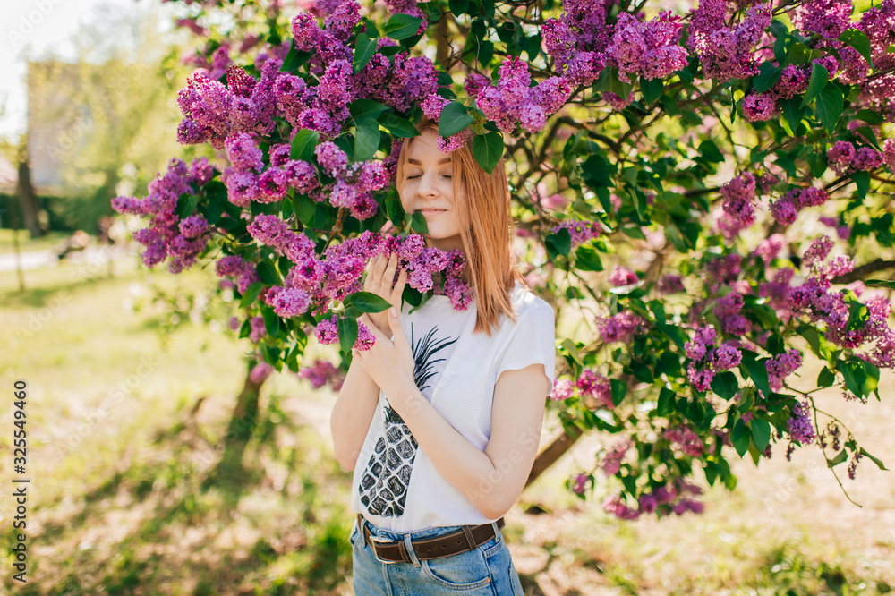 Pretty young female with red hair and fair skin smells lilac in the garden with closed eyes Pretty young female with red hair and fair skin smells lilac in the garden with closed eyes