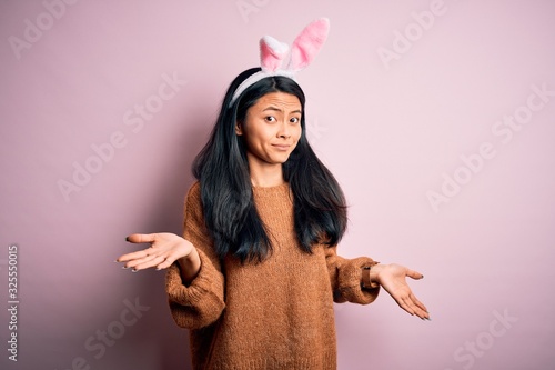 Young beautiful chinese woman wearing bunny ears standing over isolated pink background clueless and confused expression with arms and hands raised. Doubt concept.