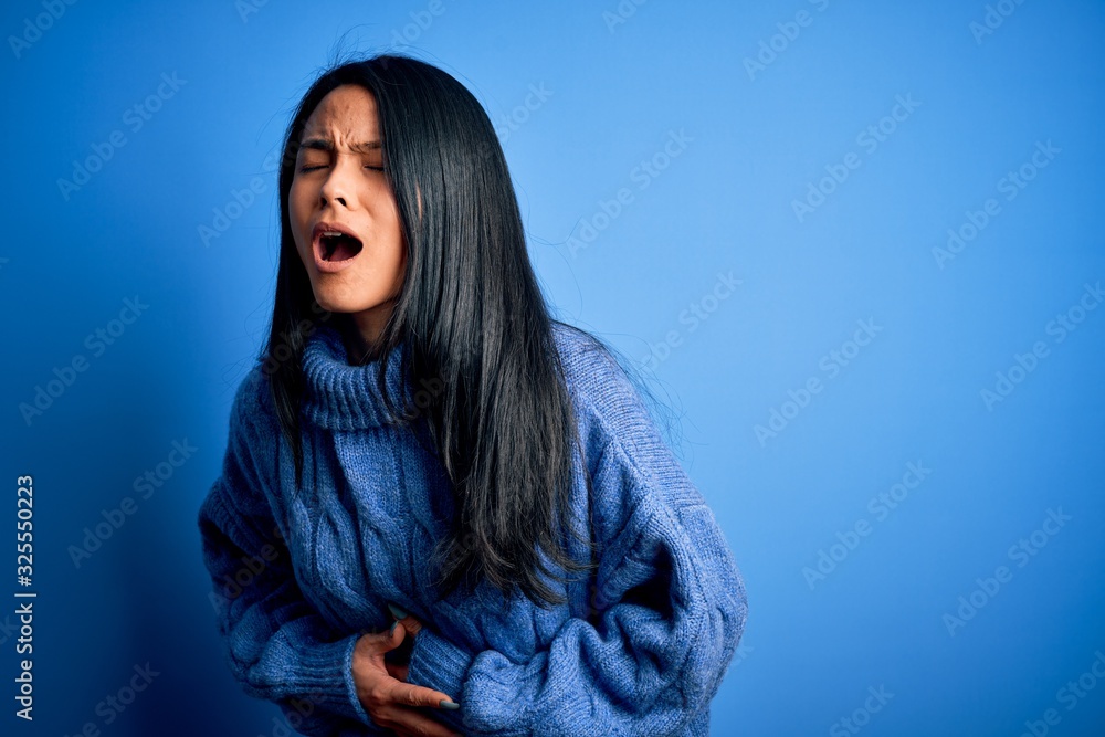 Young beautiful chinese woman wearing casual sweater over isolated blue background with hand on stomach because nausea, painful disease feeling unwell. Ache concept. Young beautiful chinese woman wearing casual sweater over isolated blue background with hand on stomach because nausea, painful disease feeling unwell. Ache concept.