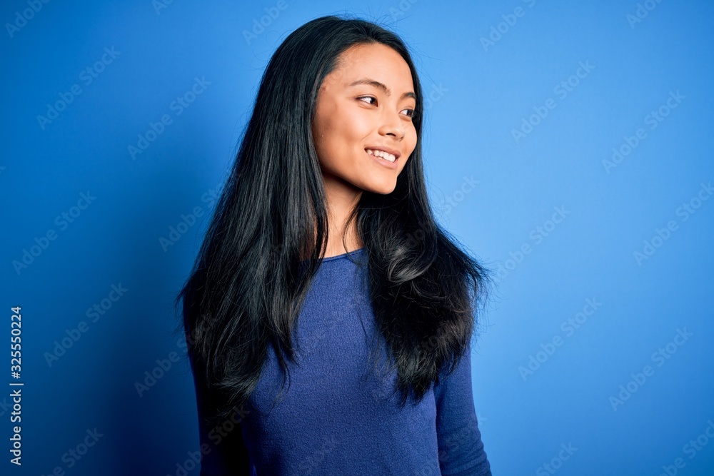 Young beautiful chinese woman wearing casual t-shirt over isolated blue background looking away to side with smile on face, natural expression. Laughing confident. Young beautiful chinese woman wearing casual t-shirt over isolated blue background looking away to side with smile on face, natural expression. Laughing confident.