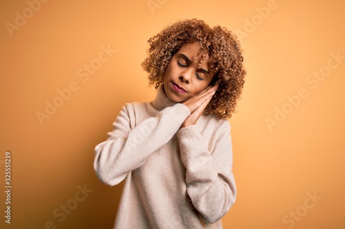 Young beautiful african american woman wearing turtleneck sweater over yellow background sleeping tired dreaming and posing with hands together while smiling with closed eyes. © Krakenimages.com