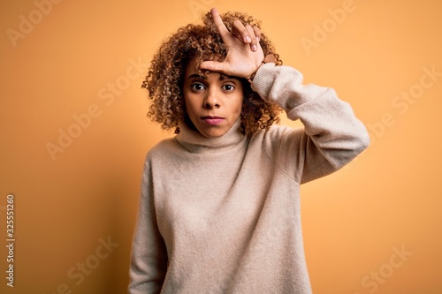 Young beautiful african american woman wearing turtleneck sweater over yellow background making fun of people with fingers on forehead doing loser gesture mocking and insulting. © Krakenimages.com