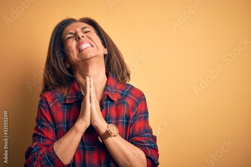 Middle age beautiful woman wearing casual shirt standing over isolated yellow background begging and praying with hands together with hope expression on face very emotional and worried. Begging.
