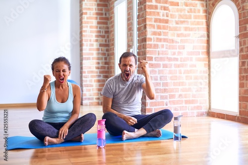 Middle age sporty couple sitting on mat doing stretching yoga exercise at gym angry and mad raising fist frustrated and furious while shouting with anger. Rage and aggressive concept.