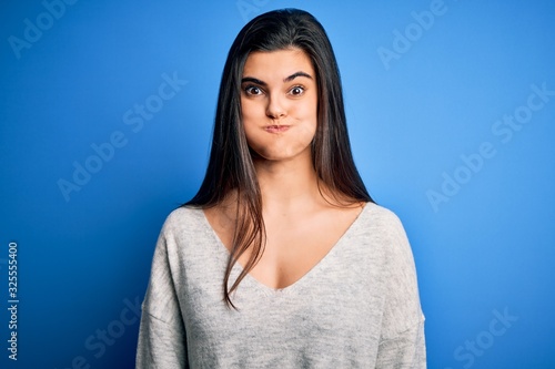 Young beautiful brunette woman wearing casual sweater standing over blue background puffing cheeks with funny face. Mouth inflated with air, crazy expression.