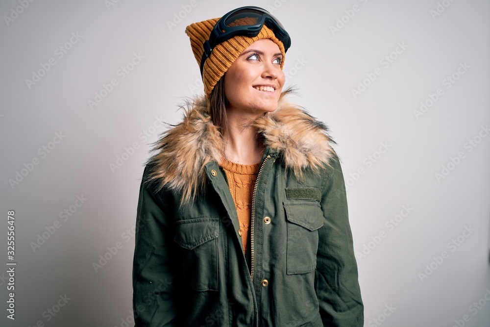 Young beautiful skier woman with blue eyes wearing snow sportswear and ski goggles looking away to side with smile on face, natural expression. Laughing confident.