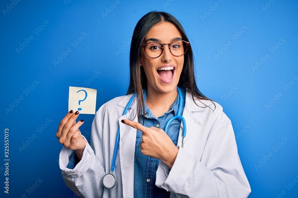 Young beautiful brunette doctor woman holding paper with question mark symbol message very happy pointing with hand and finger