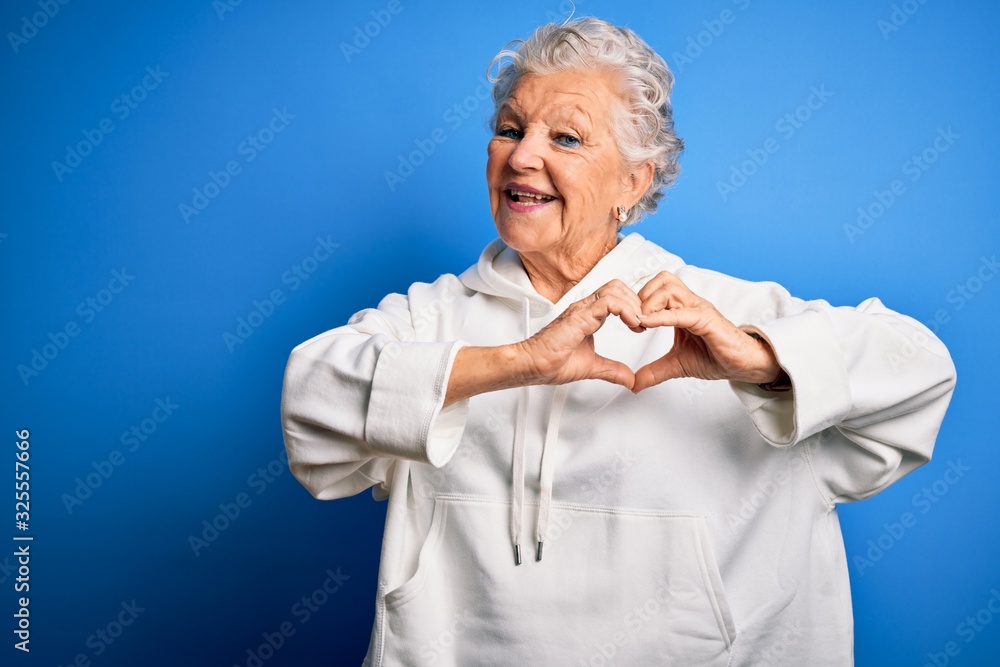 Senior beautiful sporty woman wearing white sweatshirt over isolated blue background smiling in love showing heart symbol and shape with hands. Romantic concept. Senior beautiful sporty woman wearing white sweatshirt over isolated blue background smiling in love showing heart symbol and shape with hands. Romantic concept.
