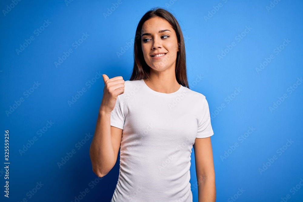 Young beautiful brunette woman wearing white casual t-shirt standing over blue background smiling with happy face looking and pointing to the side with thumb up. Young beautiful brunette woman wearing white casual t-shirt standing over blue background smiling with happy face looking and pointing to the side with thumb up.