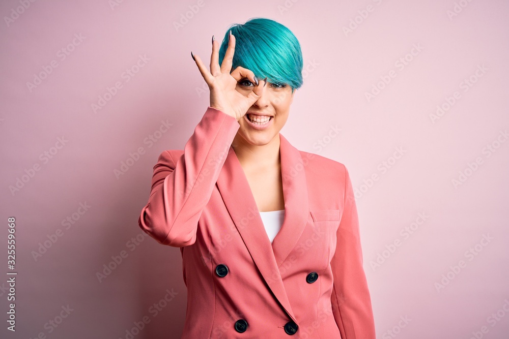Young beautiful businesswoman with blue fashion hair wearing jacket over pink background doing ok gesture with hand smiling, eye looking through fingers with happy face.