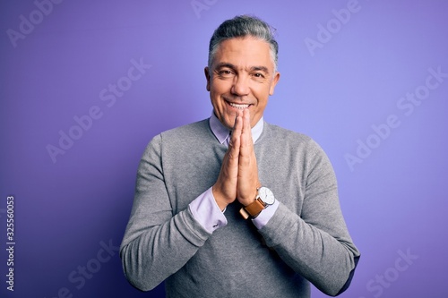 Middle age handsome grey-haired man wearing elegant sweater over purple background praying with hands together asking for forgiveness smiling confident.