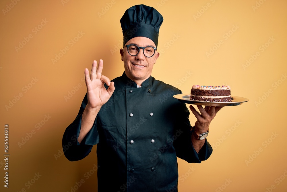 Middle age handsome grey-haired baker man wearing cooker uniform and hat holding cake doing ok sign with fingers, excellent symbol Middle age handsome grey-haired baker man wearing cooker uniform and hat holding cake doing ok sign with fingers, excellent symbol
