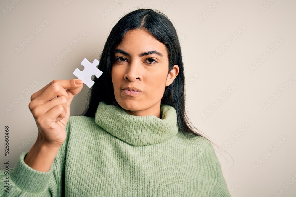 Young beautiful hispanic woman holding puzzle piece as cooperation solution on business with a confident expression on smart face thinking serious Young beautiful hispanic woman holding puzzle piece as cooperation solution on business with a confident expression on smart face thinking serious