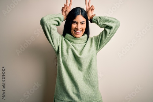 Young beautiful hispanic woman wearing green winter sweater over isolated background Posing funny and crazy with fingers on head as bunny ears, smiling cheerful © Krakenimages.com
