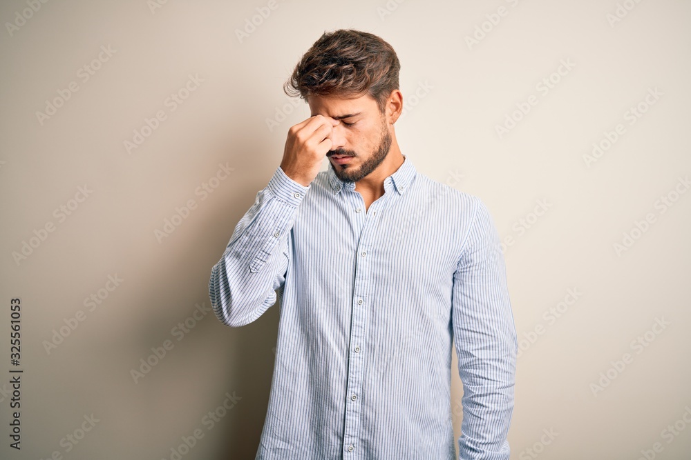 Young handsome man with beard wearing striped shirt standing over white background tired rubbing nose and eyes feeling fatigue and headache. Stress and frustration concept.