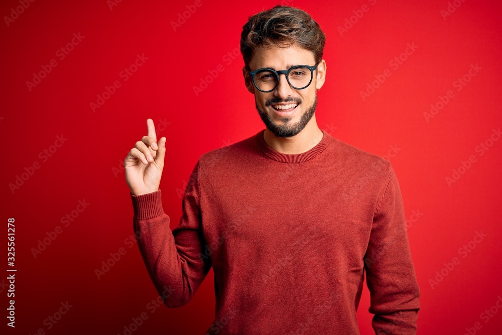 Naklejka premium Young handsome man with beard wearing glasses and sweater standing over red background with a big smile on face, pointing with hand finger to the side looking at the camera.