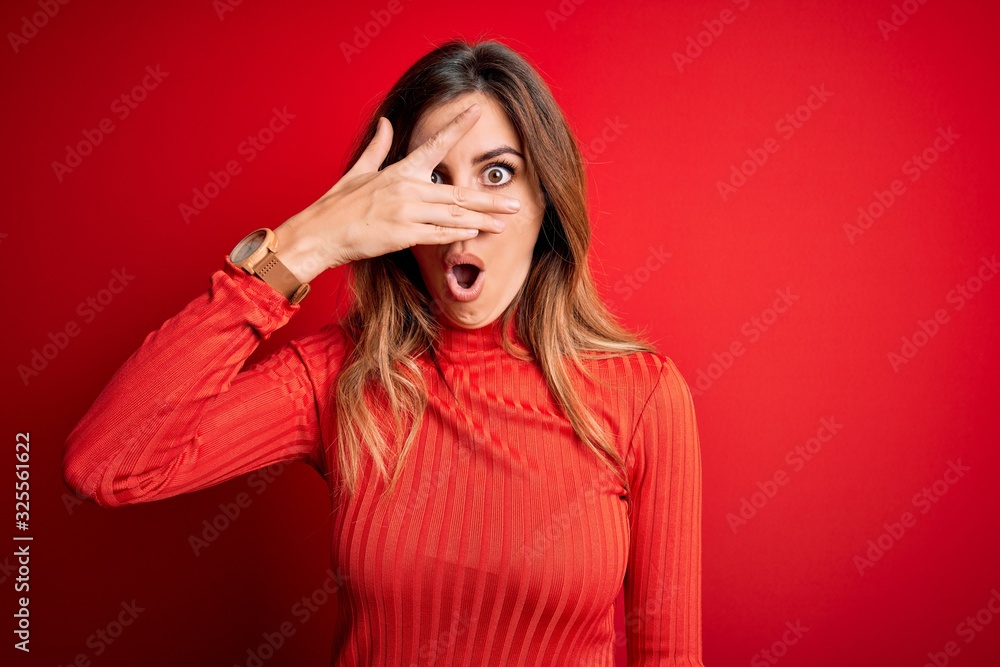 Young beautiful brunette woman wearing casual turtleneck sweater over red background peeking in shock covering face and eyes with hand, looking through fingers with embarrassed expression. Young beautiful brunette woman wearing casual turtleneck sweater over red background peeking in shock covering face and eyes with hand, looking through fingers with embarrassed expression.