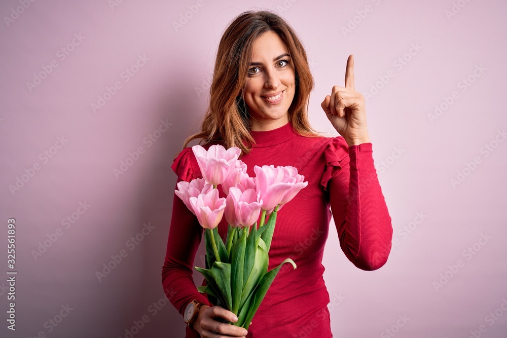 Young beautiful brunette woman holding bouquet of pink tulips over isolated background pointing finger up with successful idea. Exited and happy. Number one. Young beautiful brunette woman holding bouquet of pink tulips over isolated background pointing finger up with successful idea. Exited and happy. Number one.