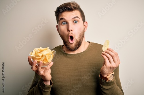 Young blond man with beard and blue eyes holding bowl with unhealthy potatoes chips scared in shock with a surprise face  afraid and excited with fear expression