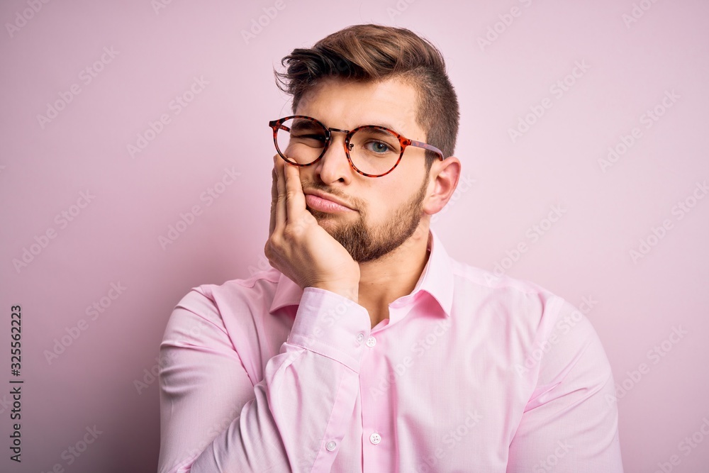 Young handsome blond man with beard and blue eyes wearing pink shirt and glasses thinking looking tired and bored with depression problems with crossed arms. Young handsome blond man with beard and blue eyes wearing pink shirt and glasses thinking looking tired and bored with depression problems with crossed arms.