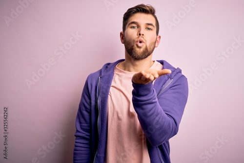 Fototapeta Naklejka Na Ścianę i Meble -  Young blond man with beard and blue eyes wearing purple sweatshirt over pink background looking at the camera blowing a kiss with hand on air being lovely and sexy. Love expression.