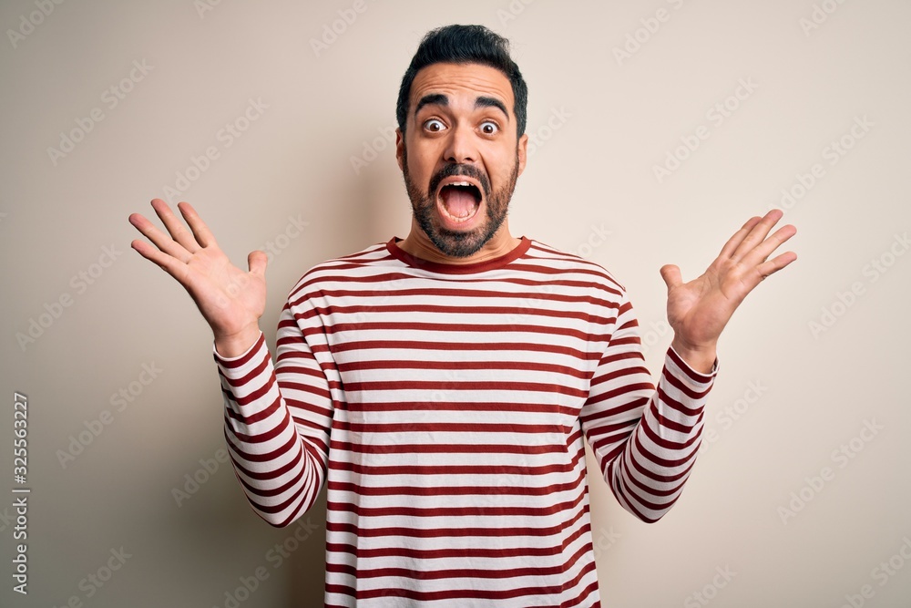 Naklejka premium Young handsome man with beard wearing casual striped t-shirt standing over white background celebrating crazy and amazed for success with arms raised and open eyes screaming excited. Winner concept