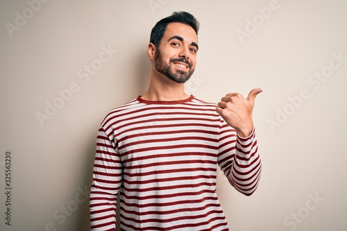 Young handsome man with beard wearing casual striped t-shirt standing over white background smiling with happy face looking and pointing to the side with thumb up.