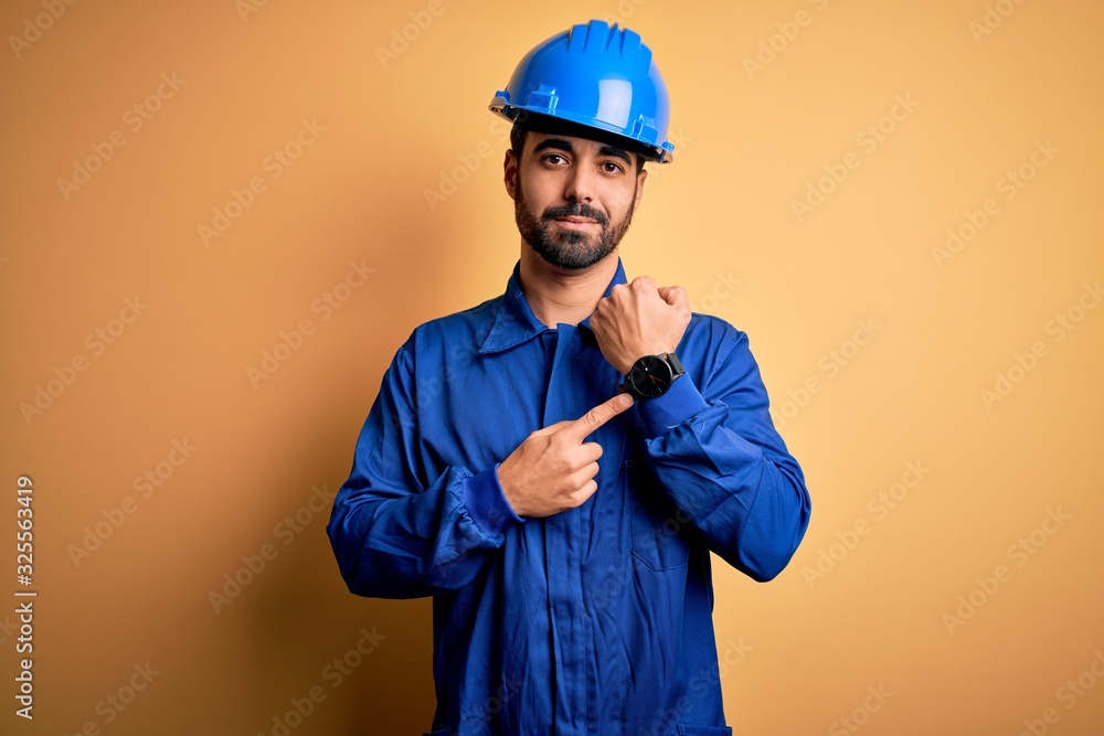 Mechanic man with beard wearing blue uniform and safety helmet over yellow background In hurry pointing to watch time, impatience, looking at the camera with relaxed expression
