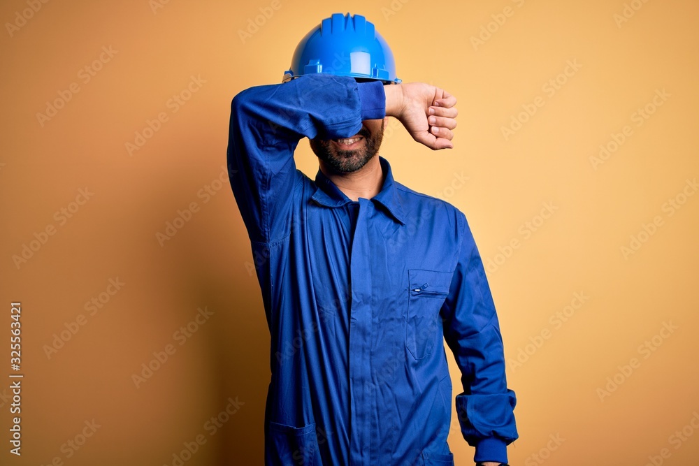 Mechanic man with beard wearing blue uniform and safety helmet over yellow background covering eyes with arm smiling cheerful and funny. Blind concept.