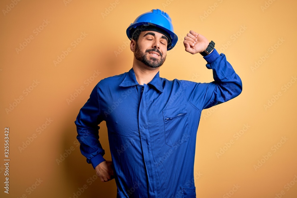 Mechanic man with beard wearing blue uniform and safety helmet over yellow background stretching back, tired and relaxed, sleepy and yawning for early morning