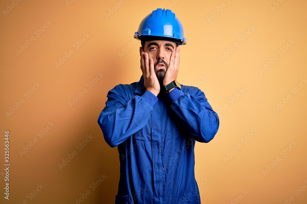 Mechanic man with beard wearing blue uniform and safety helmet over yellow background Tired hands covering face, depression and sadness, upset and irritated for problem